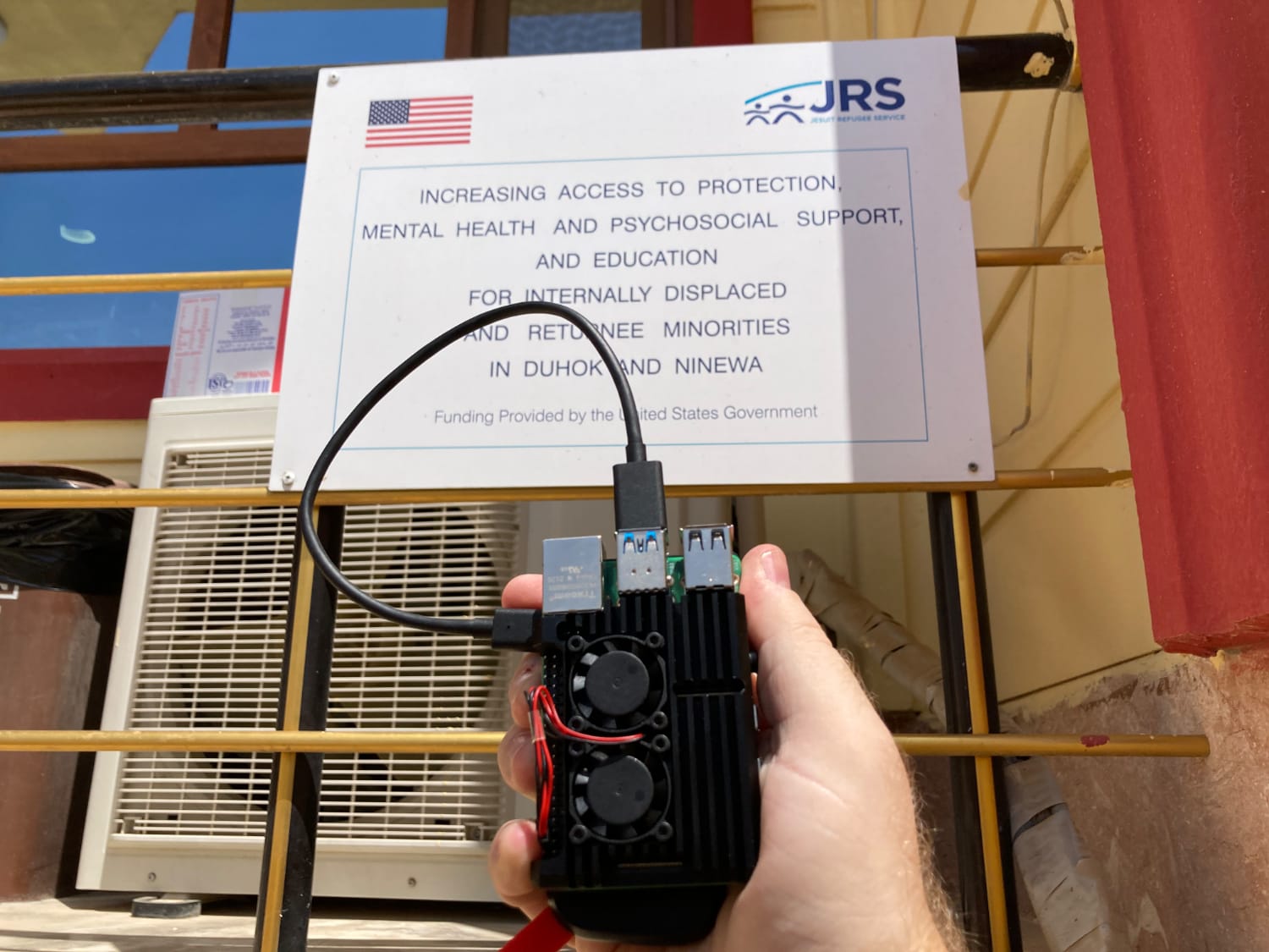 An assembled IIAB unit being held in front of a sign for the JRS Duhok office; the sign explains the mission and funding of the project there